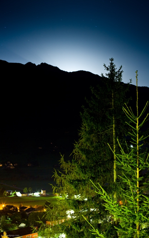 A night photograph of the village of Matrei, taken from Hotel Goldried. Various lights in the valley below illuminate the houses and fields, while moonlight casts a white glow behind the mountain in the background.